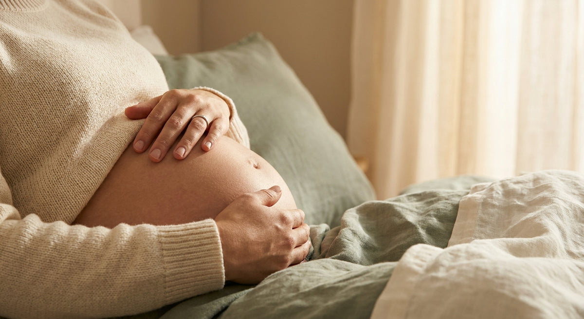 A pregnant woman gently cradling her bare belly with both hands in soft, warm natural light against a neutral background.